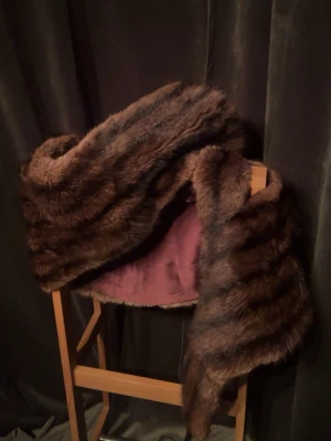 Vintage 60-tals mink – halsduk eller cape. -  Elegant mink från 1960-talet i mycket fint skick, med naturlig lyster och klassiskt randat mönster. Insidan är fodrad i burgundy satin för en lyxig känsla. Kan bäras som halsduk eller cape, och har diskreta hållare för enkel fästning över jacka eller kappa. Perfekt för vintageälskare, stylister eller dig som uppskattar äkta kvalitet. Mått: 150×30 cm.