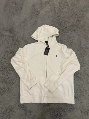 Vit hoodie från Polo Ralph Lauren - Snygg vit hoodie från Polo Ralph Lauren med dragkedja framtill och klassisk blå logga broderad på bröstet. Hoodien har huva och två fickor fram. Tillverkad i mjukt bomullsmaterial som känns skönt mot huden. Perfekt för en avslappnad och stilren look. Den är XL i barn storlek så skulle nog säga den passar någon som bär S. ⚠️tröjan har använts 2 gånger, den har prislapp, påse och qrscan⚠️
