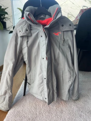 Grå vindjacka från Hollister - Snygg grå vindjacka från Hollister med röd huva och logga på bröstet. Jackan har dragkedja, knappar framtill och två fickor. Insidan av huvan är fodrad i rött tyg för extra kontrast. Perfekt för blåsiga dagar och enkel att matcha.