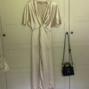 Champagnefärgad långklänning i satin - Elegant långklänning i skimrande champagnefärgad satin med djup v-ringning och korta, vida ärmar. Klänningen har en snygg twist-detalj framtill och ett mjukt, följsamt fall. Perfekt för dig som vill sticka ut med en lyxig och stilren look. ALDIRG använd!