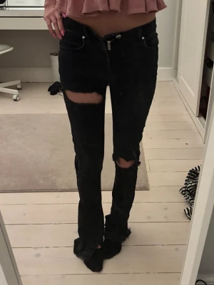 Svarta slitna jeans med hål - Säljer ett par svarta jeans med stora slitningar över knäna och raka ben. Jeansen har klassisk femficksmodell och låg midja. Perfekta för en edgy och avslappnad stil.