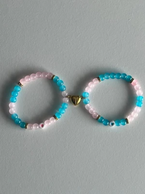 Blåt och rosa pärlarmband med magnet-hjärta - Sött armband med pärlor i ljusrosa och turkost, dekorerat med små guldfärgade detaljer. Armbandet har ett hjärtformat guldfärgat lås i mitten som binder ihop två ringar av pärlor. Perfekt accessoar för en lekfull och färgglad stil.