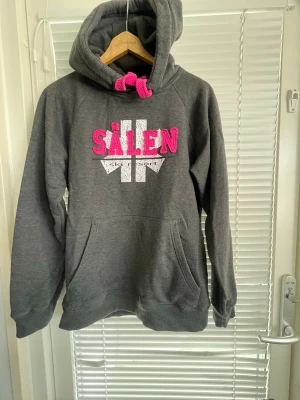 Mörkgrå Sälen hoodie med rosa detaljer - Mysig mörkgrå hoodie med stor magficka och huva. Framtill står det 'Sälen' i rosa bokstäver och vitt tryck under. Rosa dragsko i huvan och liten rosa logga bak på huvan. Perfekt för chill dagar och streetstyle. Liiiite nopprig på vissa ställen.