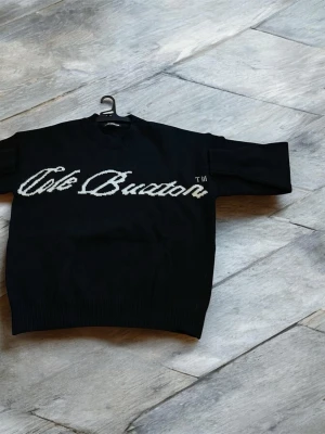 Svart stickad tröja Cole Buxton - Svart stickad tröja från Cole Buxton med vit logotyp i stor stil över bröstet. Tröjan har rund hals, långa ärmar och ribbade muddar vid ärmslut och nederkant. Perfekt för dig som gillar streetwear och vill ha en clean look med tydlig branding.