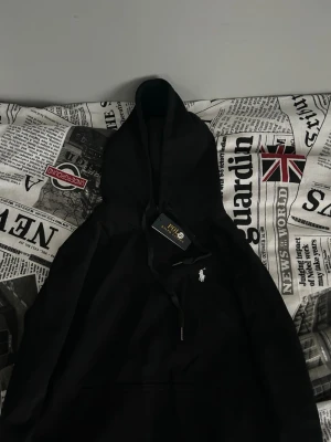 Svart hoodie från Polo Ralph Lauren - Snygg svart hoodie från Polo Ralph Lauren med klassisk huva och dragsko. Ikoniska vita loggan broderad på bröstet. Tillverkad i mjukt material som känns skönt mot huden. Perfekt för dig som gillar stilrena och tidlösa plagg. Jag beställde en hoodie i storlek S, men upplever att den är väldigt liten i storleken och motsvarar snarare en XS. Helt ny!