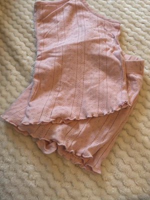 Rosa pyjamasset med volangkant - Sött pyjamasset i ljusrosa med linjer och små hålmönster. Setet består av ett linne med volangkant och matchande shorts med resår i midjan och volangdetaljer. Perfekt för mysiga kvällar hemma. 