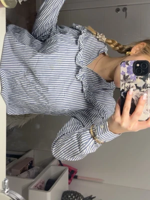 Randig blus med volang från Xoul Edition - Säljer en blå- och vitrandig blus från Xoul Edition i storlek 36. Blusen har långa ärmar, volangdetaljer längs ringningen och knäppning framtill. Tillverkad i ett lätt och luftigt material som passar perfekt till jeans eller kjol.