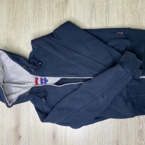 Mörkblå hoodie från GAB - Snygg mörkblå hoodie från GAB med dragkedja framtill och justerbar huva med snören. Insidan av huvan är ljusgrå och materialet känns mjukt och bekvämt. Perfekt för en avslappnad stil och enkel att matcha med jeans eller joggers.