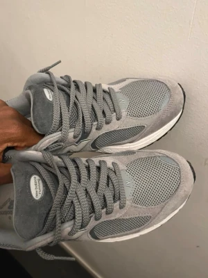 Grå New Balance 990v5 sneakers - Snygga grå New Balance 990v5 sneakers med ovandel i mocka och mesh för skön ventilation. Klassisk N-logga på sidan och tjock vit sula med svart yttersula. Perfekta för dig som gillar stilrena och bekväma sneakers.