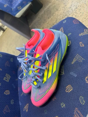Adidas f50 - Säljer ett par Adidas X Speedflow fotbollsskor i en riktigt färgstark design. Skorna har en mix av blått, rosa och neon-gult med klassiska Adidas-ränder på sidan. Ovandelen är i syntetmaterial och snörningen är blå. Perfekta för dig som vill sticka ut på planen!