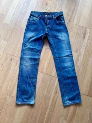 Blå raka jeans från Dr Denim - Snygga blå jeans från Detroit (Lindex) med raka ben och klassisk femficksmodell. Jeansen har slitningar och tvättade detaljer för en cool, avslappnad look. Tillverkade i bomull med knappgylf och läderpatch bak i midjan.