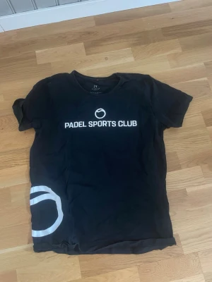 Svart Padel Sports Club t-shirt - Svart t-shirt från IN med vit text 'PADEL SPORTS CLUB' och en vit cirkeldetalj nere till vänster. Klassisk rund hals och korta ärmar. Perfekt för dig som gillar sportig stil.