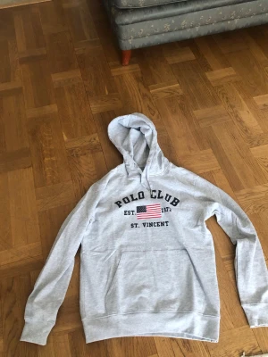 Grå hoodie från Polo Club - Snygg ljusgrå hoodie från Polo Club med tryck framtill som inkluderar en amerikansk flagga och texten 'EST. 1972 ST. VINCENT'. Hoodien har en stor ficka. Har knappt använt den för den va lite för stor för mig. 
