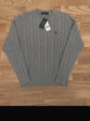 Stickad Ralph Lauren - Kabelstickad grå tröja från Polo Ralph Lauren med rund halsringning och ribbade muddar. Klassisk marinblå logga broderad på bröstet.  Tidlös modell med broderad logga på bröstet. Helt ny med tags, aldrig använd. Kostar 650 men kan få en beige också för 1000kr tillsammans 