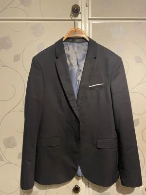 Svart kavaj från Zara - Stilren svart kavaj från Zara med två knappar framtill, klassisk krage och fickor med lock. Fodrad med ljusgrått tyg och har en vit detalj vid bröstfickan. Perfekt för en dressad look.