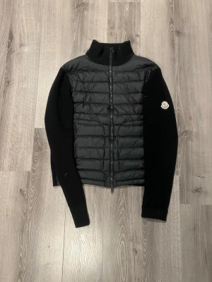 Monclear cardigan - Snygg svart dunväst från Moncler med quiltad front och stickade ärmar. Västen har hög krage, dragkedja framtill och Moncler-logga på ena ärmen. Perfekt för lager-på-lager och ger en clean look.