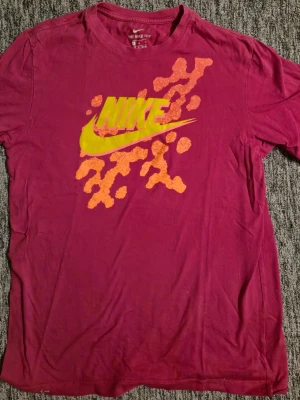 Vinröd/lila Nike t-shirt med tryck - Cool röd t-shirt från Nike med stort gult logotryck och orange grafiskt mönster på bröstet. Klassisk rund hals och korta ärmar. Perfekt för dig som gillar sportig och avslappnad stil. Storlek medium finns i hässelby eller skickas
