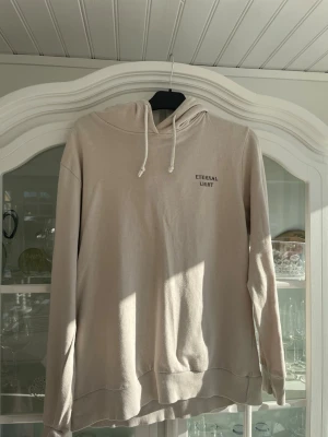 Beige hoodie från The Black Label - Snygg beige hoodie från The Black Label Collection i storlek M. Hoodien har dragsko i huvan och trycket 'ETERNAL LIGHT' på bröstet. Tillverkad i mjukt bomullsmaterial och har ribbade muddar vid ärmslut och nederkant. Perfekt för en avslappnad och stilren look.