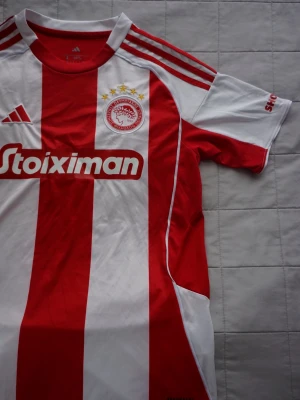 Olympiakos | matchtröja  - Snygg Olympiakos matchtröja från Adidas i rött och vitt med klassiska breda ränder. Tröjan har klubbmärke med fem stjärnor, Stoiximan som sponsortryck och Adidas logga. Tillverkad i lätt och ventilerande material, perfekt för fotboll eller träning. Helt ny och oanvänd, köpt i Aten. 