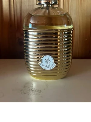 Moncler Pour Femme parfym guld - Exklusiv parfym från Moncler i en lyxig, rundad flaska med guldfärgad, räfflad yta och genomskinligt lock. Flaskan har Monclers logotyp framtill och ger ett elegant intryck. Perfekt för dig som gillar snygg design och vill ha något unikt på parfymhyllan. Skriv till mig innan köp , som ni ser är den öppnad🩵