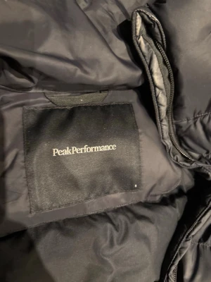 Svart dunjacka Peak Performance - Säljer en svart dunjacka från Peak Performance med huva och dragkedja framtill. Jackan har två sidofickor med dragkedja och diskret logga på bröstet. Perfekt för kalla vinterdagar och riktigt skön att bära.
