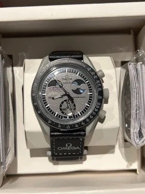 Omega x Swatch Speedmaster Moonwatch - Stilren Omega x Swatch Speedmaster Moonwatch med grå urtavla, svarta och vita detaljer samt svart armband med Omega-logga. Klockan har kronograf och tachymeter, och en unik design inspirerad av månen.