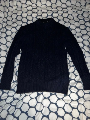 Mörkblå kabelstickad tröja - Snygg mörkblå kabelstickad tröja med rund hals och ribbade muddar vid ärmslut och nederkant. Klassisk modell som passar perfekt till jeans eller över en skjorta. Tröjan har ett diskret mönster och är gjord i mjukt material.