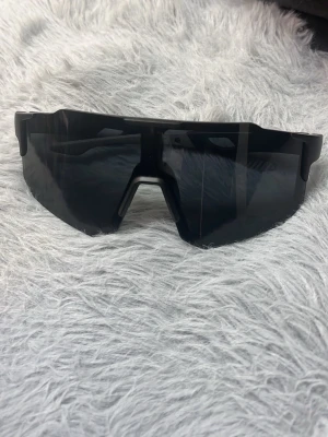 Svarta sportiga solglasögon - Säljer ett par svarta solglasögon med sportig och futuristisk design. Glasögonen har breda, rektangulära linser och markerad båge i plast. Perfekta för dig som gillar en modern look och vill sticka ut.