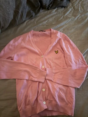 Ljusrosa kofta från Lyle & Scott - Säljer en ljusrosa kofta från Lyle & Scott med klassisk gul logga på bröstet. Koftan har v-ringning, vita knappar och ribbade muddar vid ärmslut och nederkant. Perfekt för lager på lager och enkel att matcha.