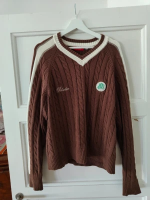 Brun kabelstickad tröja Pellador - Brun kabelstickad tröja från Pellador med vita detaljer längs axlar och ärmar. V-ringad halsringning med vit kant, broderad logga på bröstet och texten 'Pellador' på ryggen. Långa ärmar och ribbade muddar. Perfekt för dig som gillar retrostil.