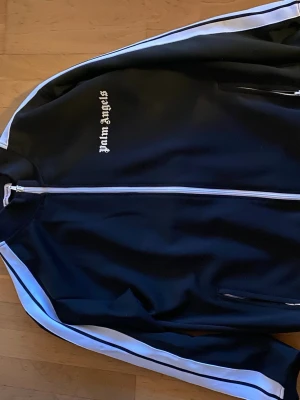 Palm Angels svart vindjacka - Snygg svart vindjacka från Palm Angels med vita ränder längs ärmarna och vit dragkedja. Jackan har ståkrage och logga broderad på bröstet i vit text. Två fickor med dragkedja framtill. Passar M och L. Använd 2 gånger