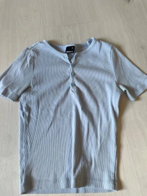 Ljusblå ribbad topp från Gina Tricot - Supersöt ljusblå ribbad topp från Gina Tricot med korta ärmar och rund halsringning. Toppen har tre knappar framtill som en snygg detalj. Perfekt att styla med jeans eller kjol för en fräsch look.
