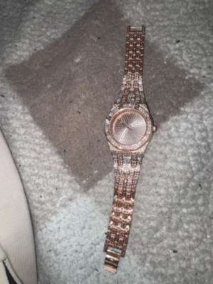 Roséguld klocka med stenar - Snygg klocka i roséguld med glittrande stenar på både urtavla och armband. Klockan har ett metallarmband med länkdesign och rund urtavla utan siffror. Perfekt accessoar för dig som gillar bling och vill sticka ut.