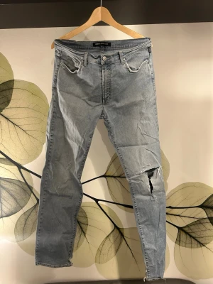 Ljusblå Slitna Versace Jeans - Säljer ett par ljusblå jeans från Versace med raka ben och hög midja. Jeansen har slitningar och ett stort hål på ena benet samt råa kanter vid bensluten. Klassisk femficksmodell och knappgylf. Perfekta för en avslappnad streetstil.