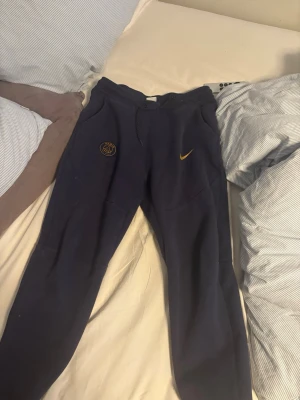 Mörkblå Nike PSG mjukisbyxor - Säljer ett par mörkblå mjukisbyxor från Nike med PSG-logga och gul Nike swoosh på vänster ben. Byxorna har dragsko i midjan och är tillverkade i mjukt material, perfekta för chill dagar. Snygg och sportig look som passar till hoodie eller t-shirt.