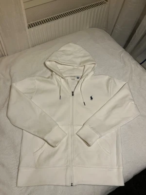Vit hoodie från Ralph Lauren - Snygg vit hoodie från Ralph Lauren med dragkedja framtill och klassisk huva. Tröjan har två fickor fram och den ikoniska Polo-loggan broderad på bröstet. Tillverkad i mjuk bomull för en skön och avslappnad stil. Perfekt till chill dagar eller när du vill vara casual.