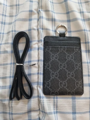 Svart korthållare med band - Snygg svart korthållare med grått monogrammönster och ring för nyckelband. Tillverkad i konstläder och har plats för flera kort. Perfekt att ha runt halsen med det medföljande svarta bandet.