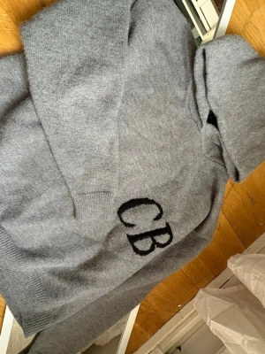 Cole Buxton - Mysig grå hoodie i mjukt stickat material med svarta bokstäverna 'CB' broderade på framsidan. Tröjan har huva och ribbade muddar vid ärmslut och nederkant. Perfekt för en chill och avslappnad stil.