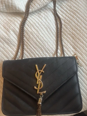 Svart YSL handväska med kedjerem - Snygg svart handväska från YSL med guldfärgad logga och dekorativ tofs framtill. Väskan har quiltat mönster och en lång kedjerem som kan bäras över axeln eller crossbody. Perfekt accessoar för att lyfta vilken outfit som helst.
