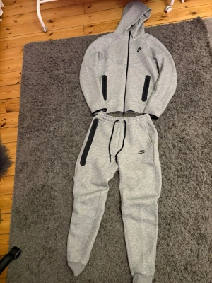 Gråa mjukisbyxor från Nike - Säljer ett par gråa mjukisbyxor från Nike med svarta detaljer och dragsko i midjan. Byxorna har muddar vid bensluten och en liten Nike-logga på vänster ben. Matchar perfekt med hoodie i samma färg för en sportig och chill look.