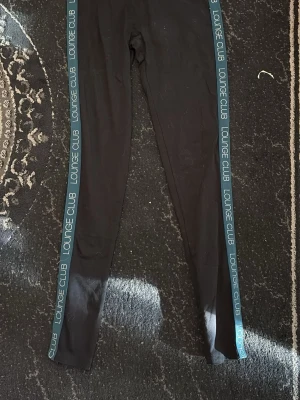 Svarta leggings med blå sidoremsor två st storlek M/S - Snygga svarta leggings med blå sidoremsor där det står 'LOUNGE CLUB' i vitt. Tight passform och stretchigt material som ger en sportig vibe. Perfekta till en avslappnad look eller när du vill vara bekväm men ändå se cool ut.