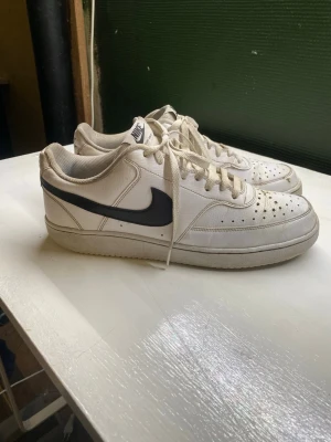 Vita Nike Air Force 1 sneakers - Klassiska vita Nike Air Force 1 sneakers med svart swoosh på sidan. Skorna har perforerad tå för ventilation, vita skosnören och en låg silhuett. Tillverkade i läder med textildetaljer och gummisula. Perfekta för en clean och sportig stil.