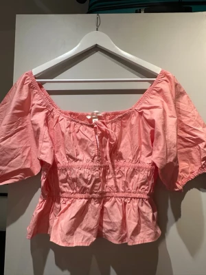 Korall blus med volang - Söt rosa offshoulder blus med puffiga korta ärmar och volang nedtill. Toppen har smock i midjan och en liten rosett framtill. Perfekt för dig som vill ha en feminin och trendig look.