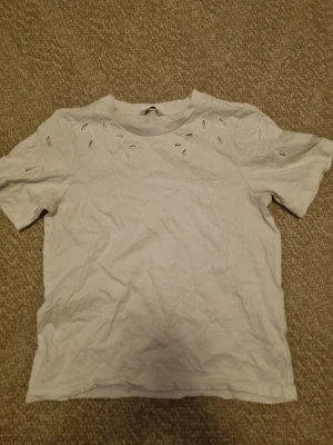 Vit t-shirt med broderade detaljer - Säljer en vit t-shirt med korta ärmar och snygga broderade detaljer samt små utskärningar vid axlar och bröst. T-shirten har en klassisk passform och är tillverkad i mjuk bomull. Perfekt för dig som gillar stilrena plagg med en twist.