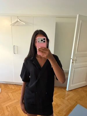 Svart kortärmad skjorta med knappar - Stilren svart skjorta med korta ärmar och dubbelknäppning framtill. Skjortan har en avslappnad passform och klassisk krage. Perfekt för dig som gillar en enkel men trendig look. Materialet ser ut att vara lätt och luftigt, troligen bomull eller linne.