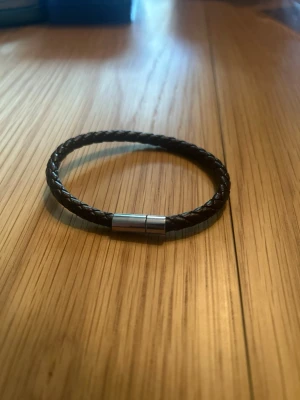 Flätat brunt läderarmband - Snyggt armband i mörkbrunt flätat läder med ett stilrent magnetlås i silverfärgad metall. Perfekt accessoar för dig som gillar en enkel men trendig look. Passar till de flesta outfits och är lätt att matcha med andra smycken.