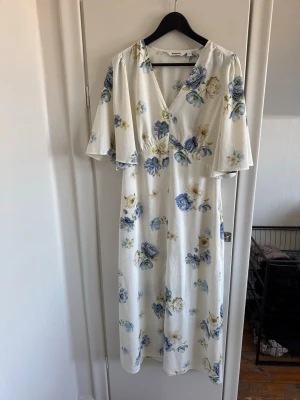 Blommig vit klänning från Kappahl - Fin vit klänning från Kappahl med blå och gula blommor. Klänningen har v-ringning, omlott-look och vida korta ärmar. Materialet känns lätt och flowy, perfekt för varma dagar. Blommönstret ger en romantisk vibe och längden är midilång.