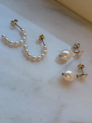 Två par örhängen med pärlor - Säljer två par örhängen i silverfärg med vita pärlor. Ett par är halvcirkelformade hoops med små pärlor, det andra paret har en större pärla som hänger från en mindre pärla. 