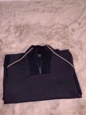 Svart ziptröja från BOSS - Snygg svart ziptröja från BOSS med diskret logga på bröstet och dragkedja framtill. Tröjan har en sportig look med kontrastfärgade detaljer längs axlarna och hög krage. Ny pris 2500 kr