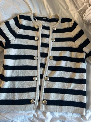 Randig kofta från BikBok XS - Snygg kofta från BikBok i beige och svart med breda horisontella ränder. Koftan har rund halsringning, dekorativa guldknappar framtill och är stickad i mjukt material. Inget fler på tröjan!